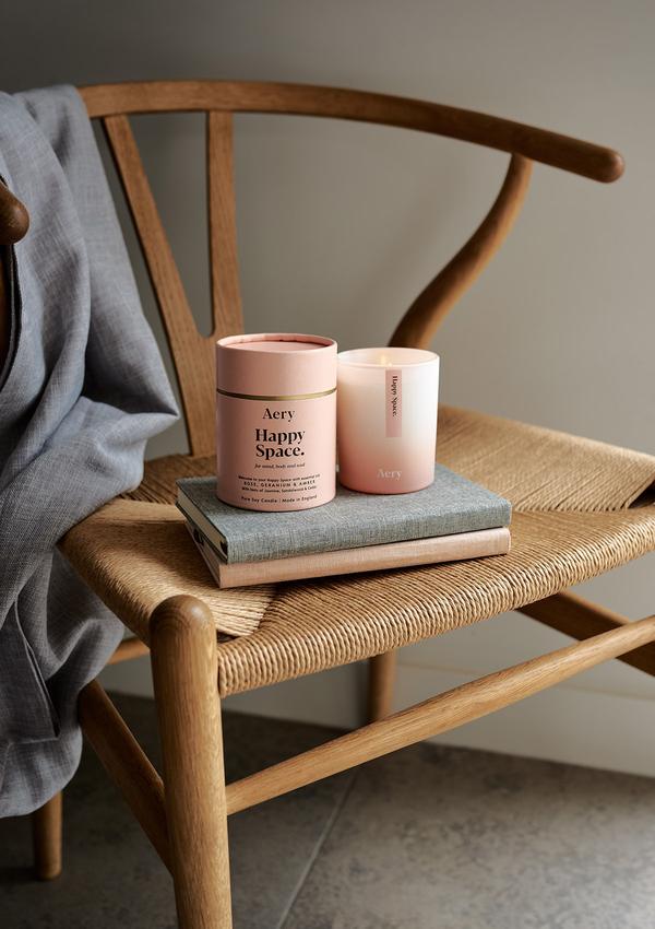 Doftljuset Happy Space från Aery Living som luktar Rose, Geranium och Amber. Produkten står på två böcker i en designer stol i trä.