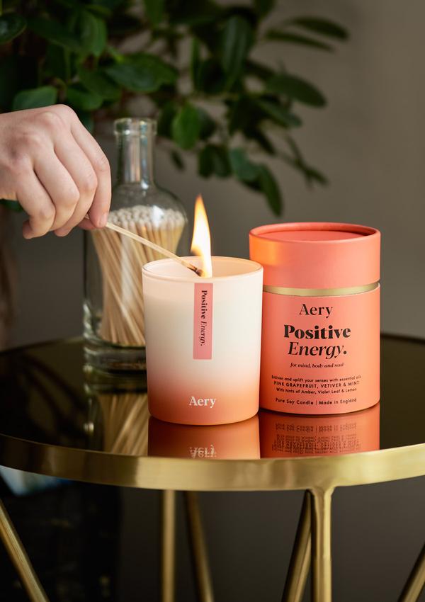 Doftljuset Positive Energy från Aery Living. Ljuset luktar Rosa grapefruit, vetiver och mint. Produkten står på ett guldbord och en person håller på att tända ljuset. 